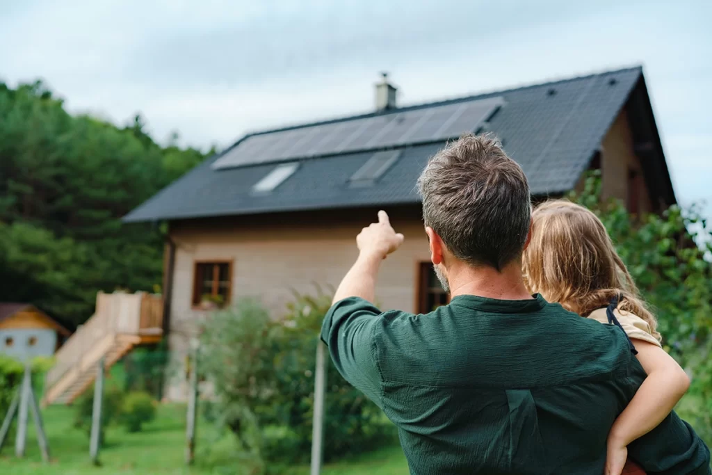 Dad holding child in arms and pointing at solar panels installed on the roof of a house