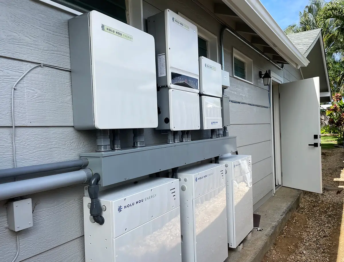 Exterior wall of a house with multiple white solar battery storage units and electrical components installed, with conduit pipes running along the wall and an open door leading inside.